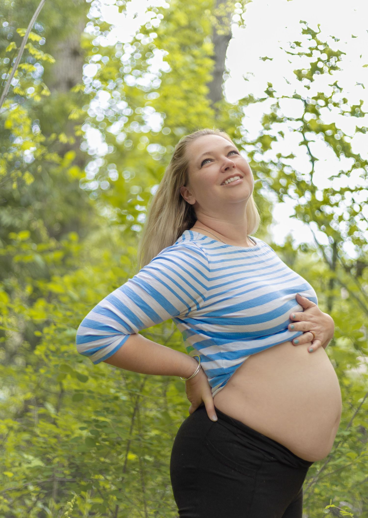 Smiling pregnant woman in blue striped shirt holding her bare belly outdoors among green trees.