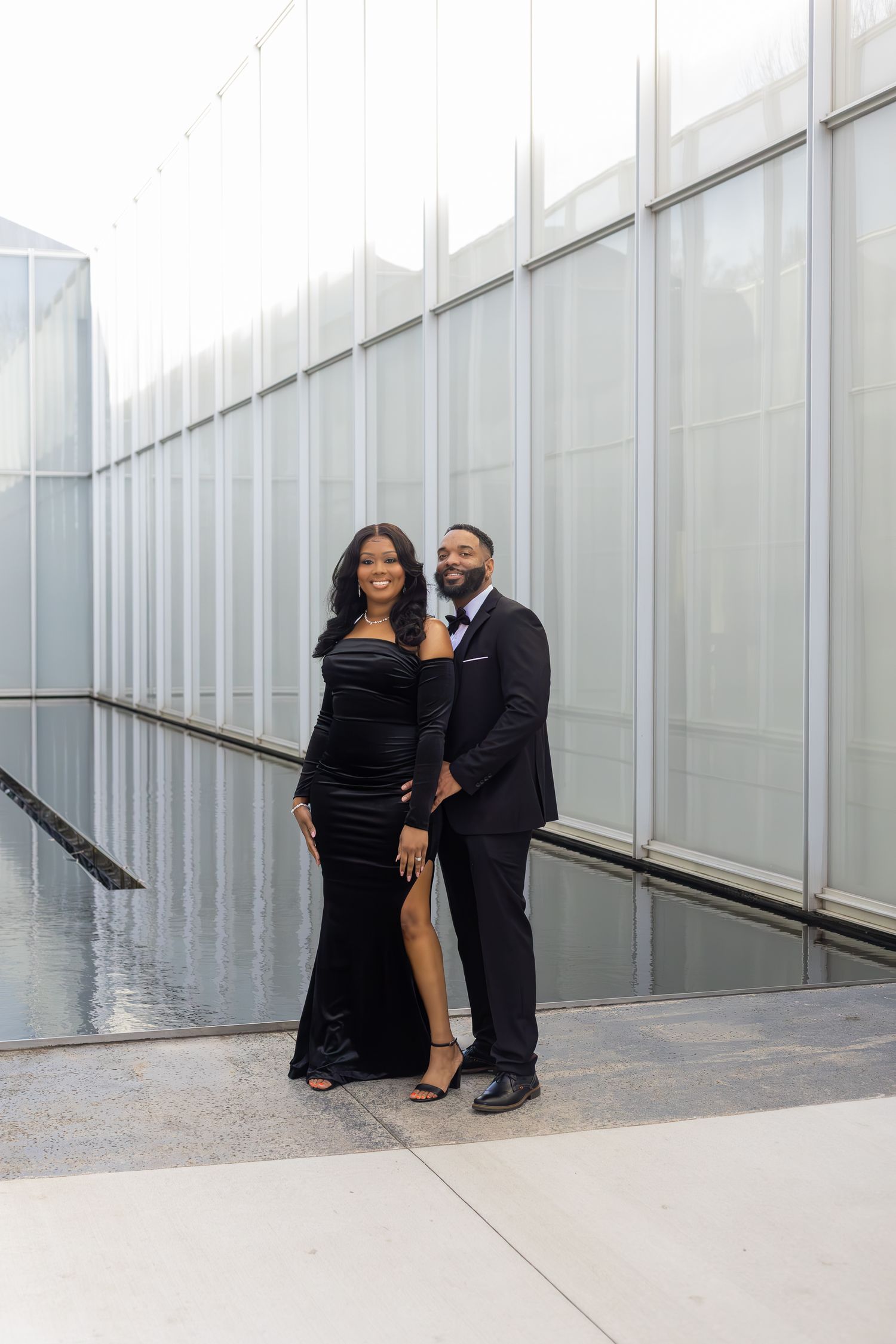A couple poses against a modern glass building wall in formal attire.