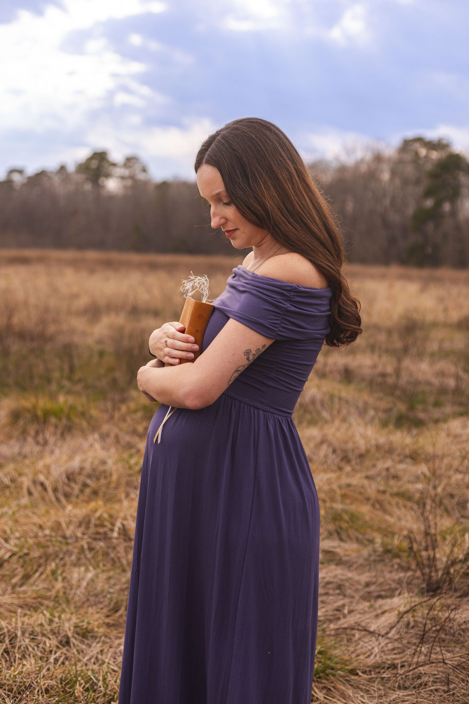 A maternity photo shoot in a field with a woman wearing a navy blue off-shoulder dress.
