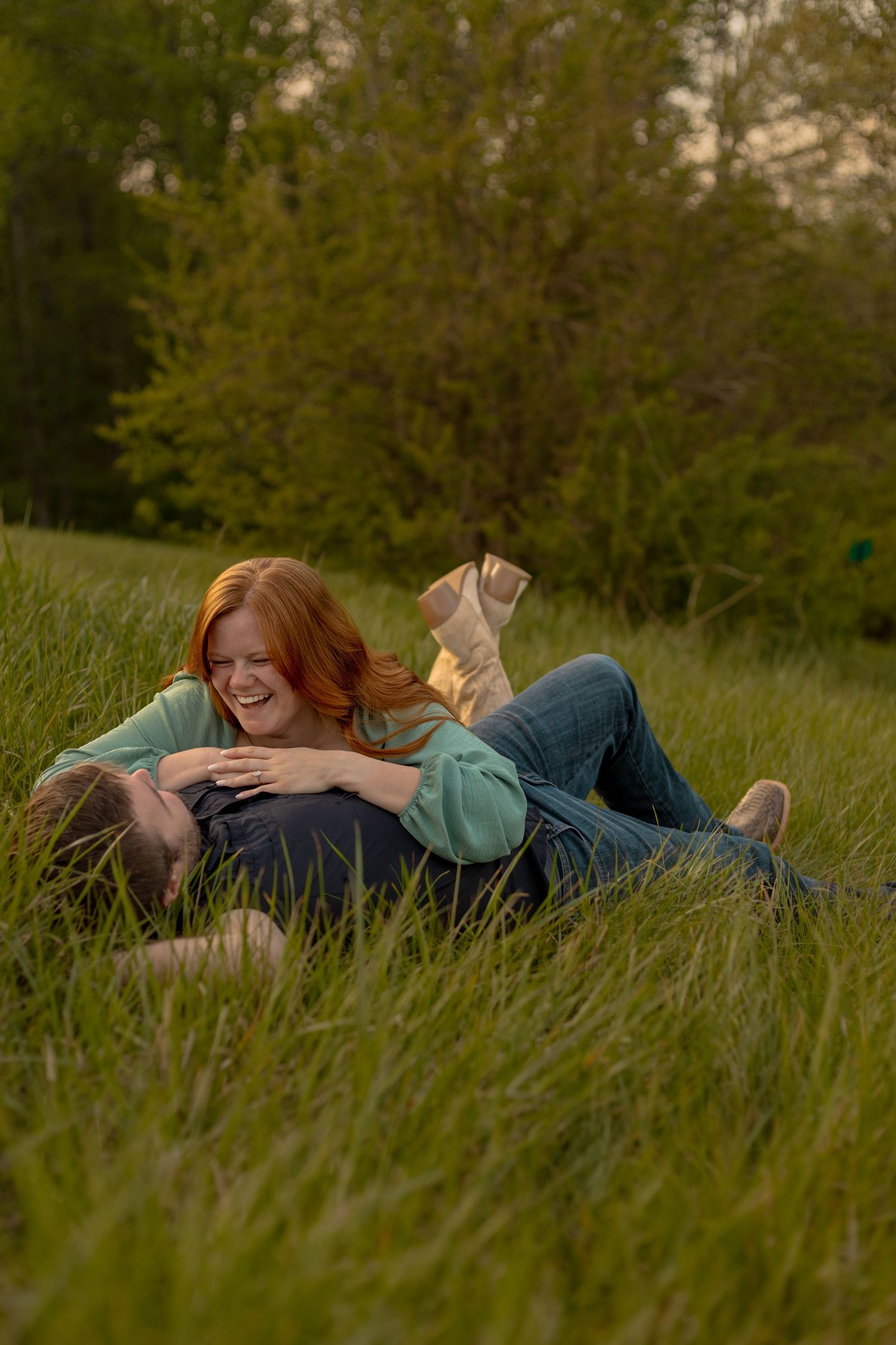 A laughing redhead woman in a green top cuddles with her partner while lying in lush green grass outdoors.