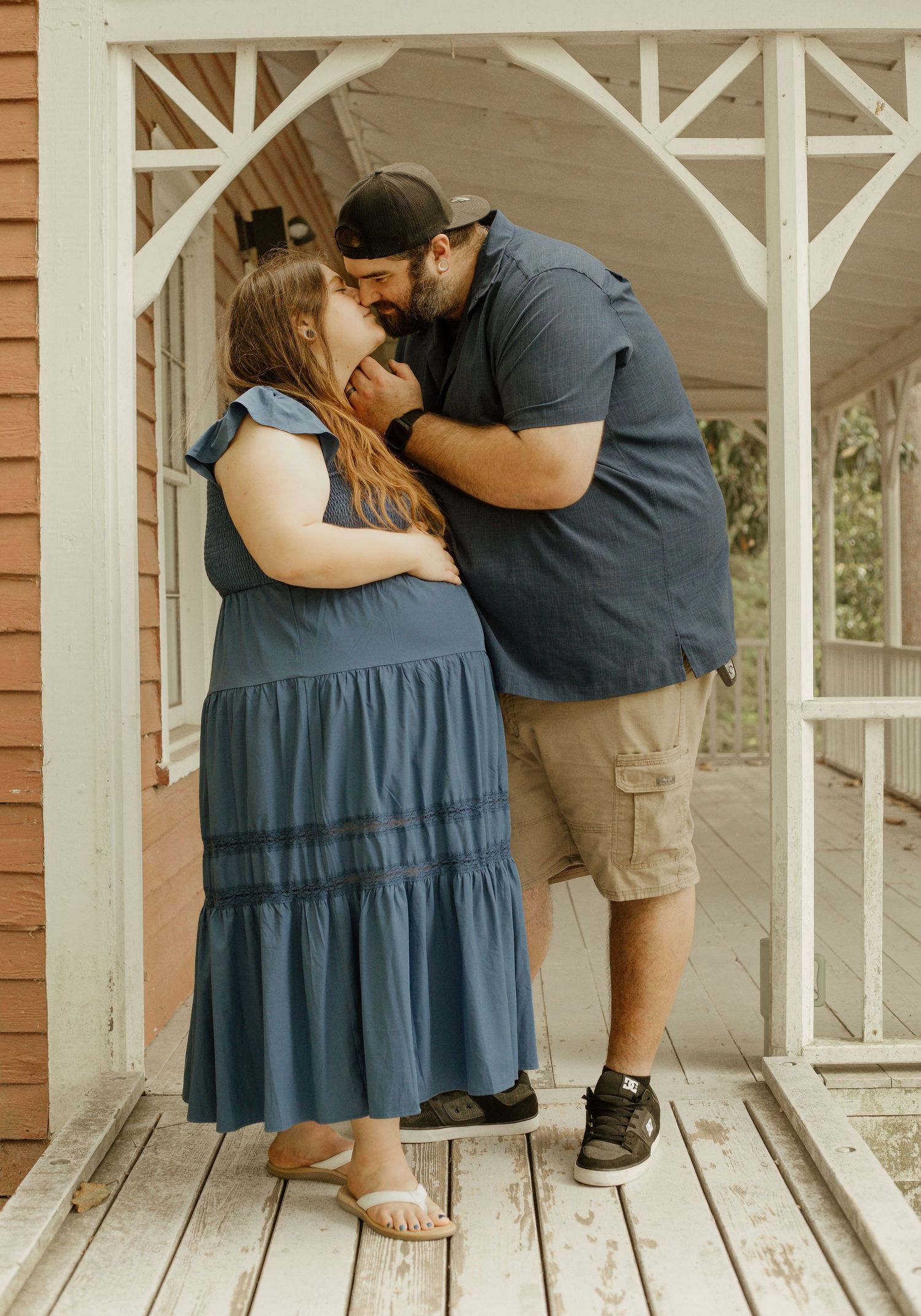 Couple sharing a tender kiss on a wooden porch, woman in blue dress, man in navy shirt and khaki shorts.