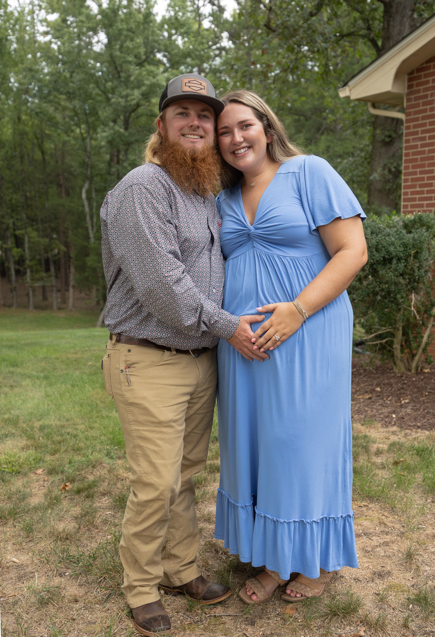 Smiling couple posing outdoors, woman in blue maternity dress holding baby bump, man in plaid shirt and cap.