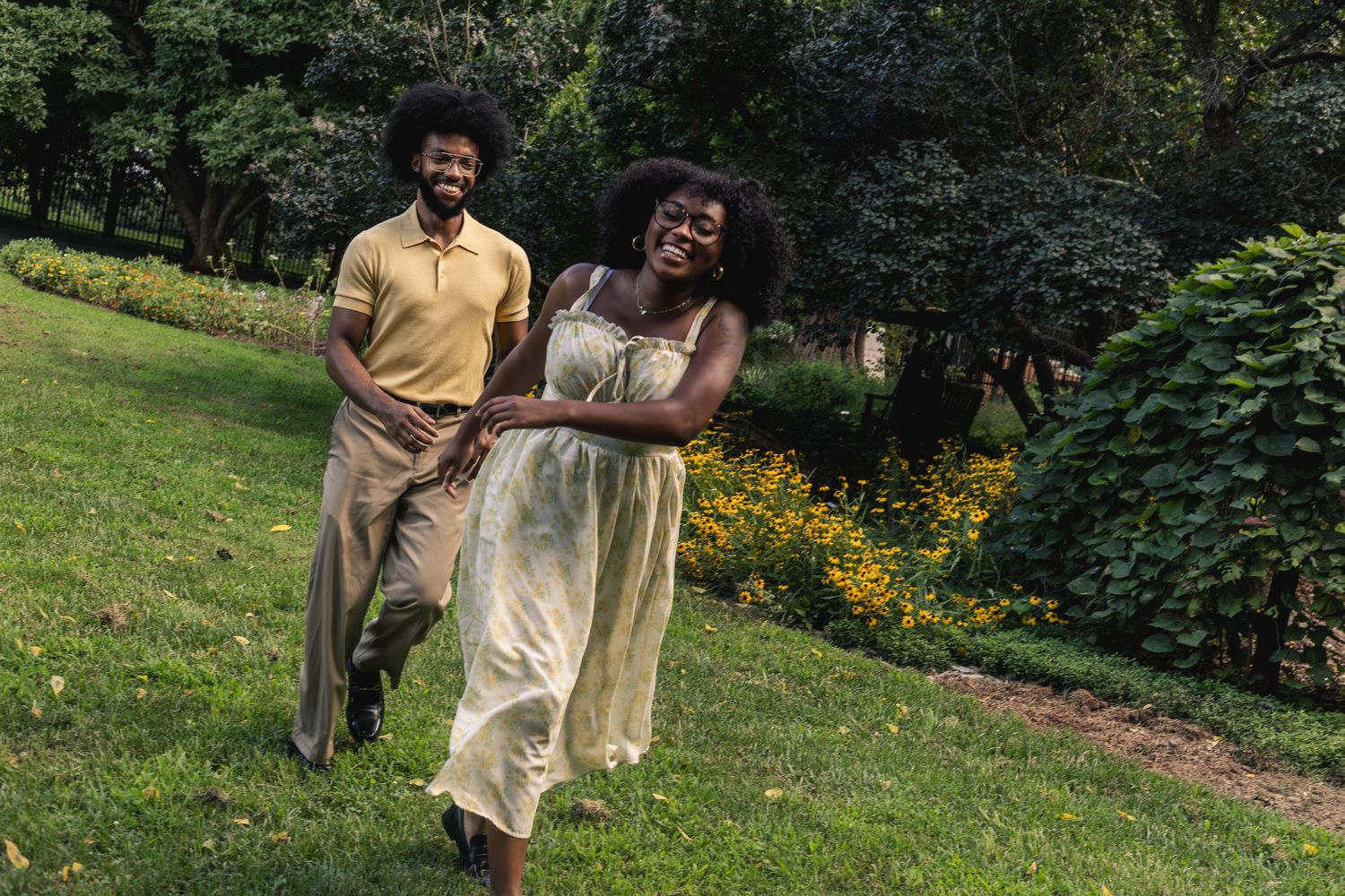 Happy Black couple holding hands running through a lush green park, woman in flowy dress, man in yellow polo.