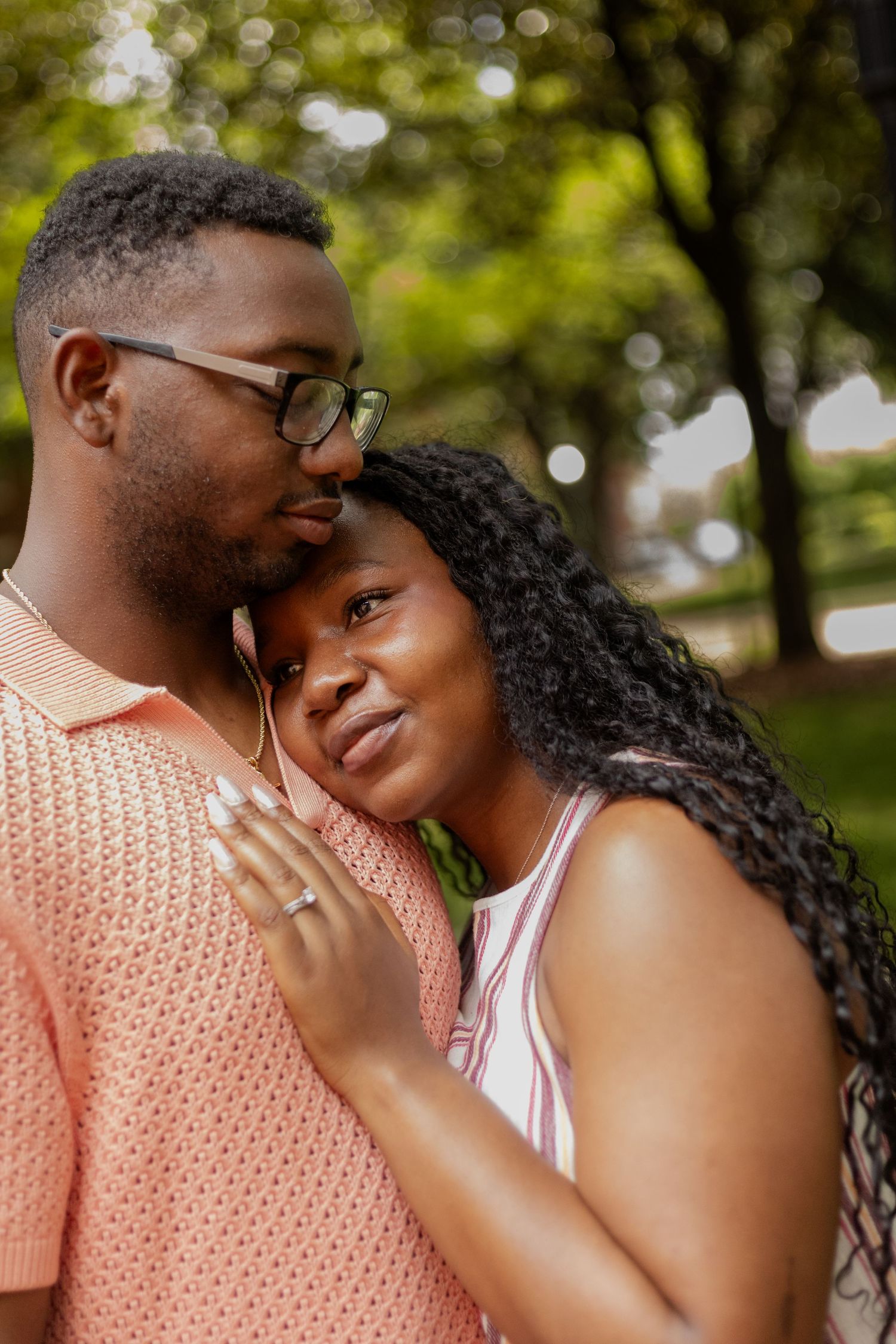 A loving Black couple shares a tender moment outdoors, with the woman resting her head on the man's chest in a lush green park.