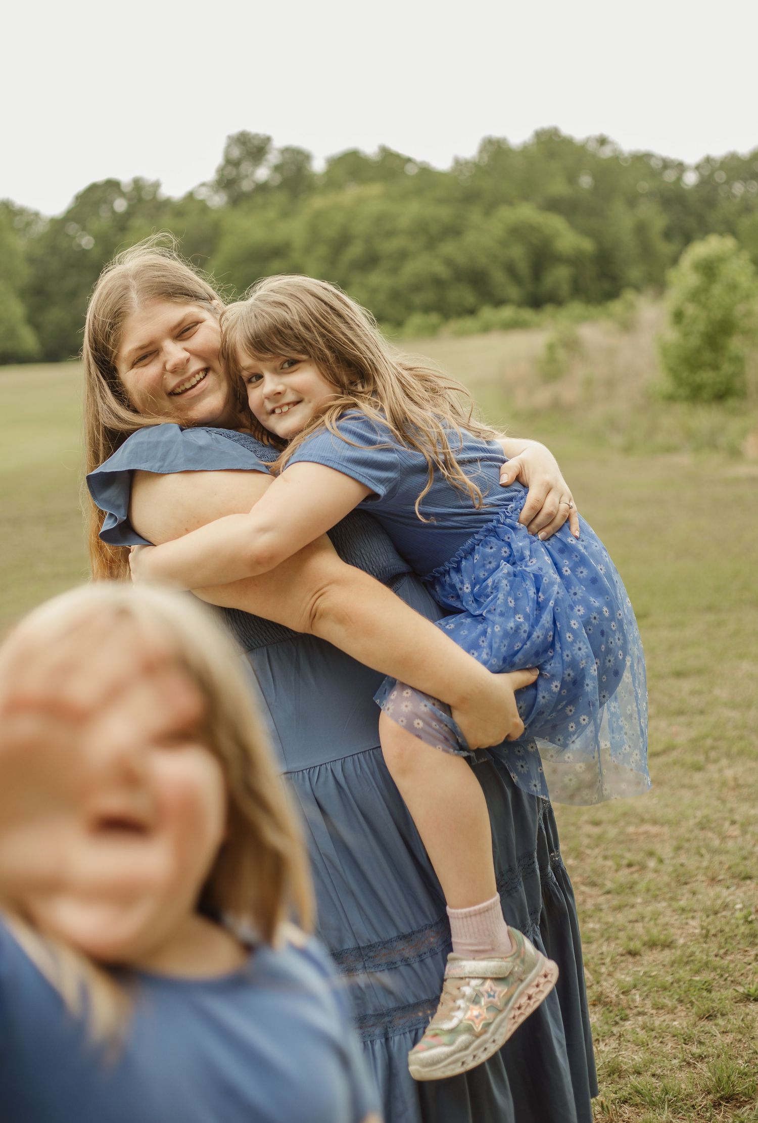 A smiling woman in a blue dress hugs a young girl outdoors in a green field, with another child in the foreground.