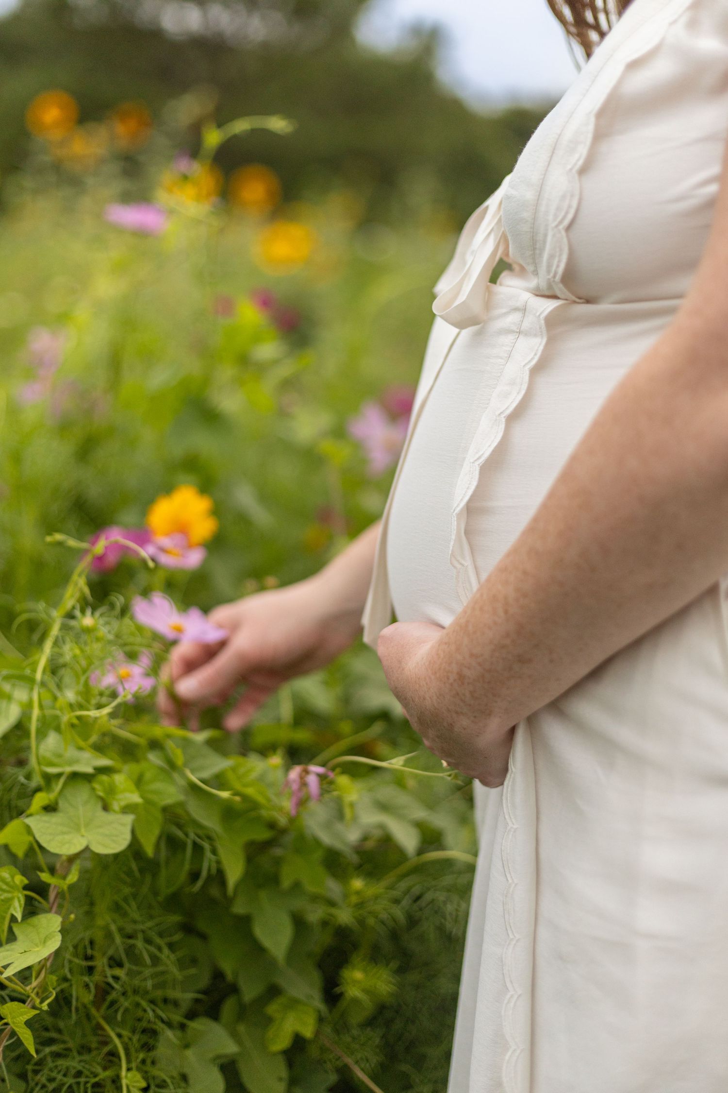 Pregnant woman in white dress touching pink flowers in a lush garden with colorful blooms.