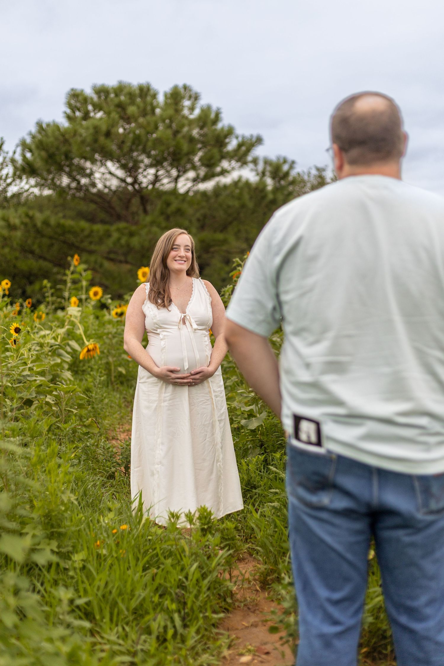 Pregnant woman in white dress holds belly in sunflower field while partner watches during maternity photo session.