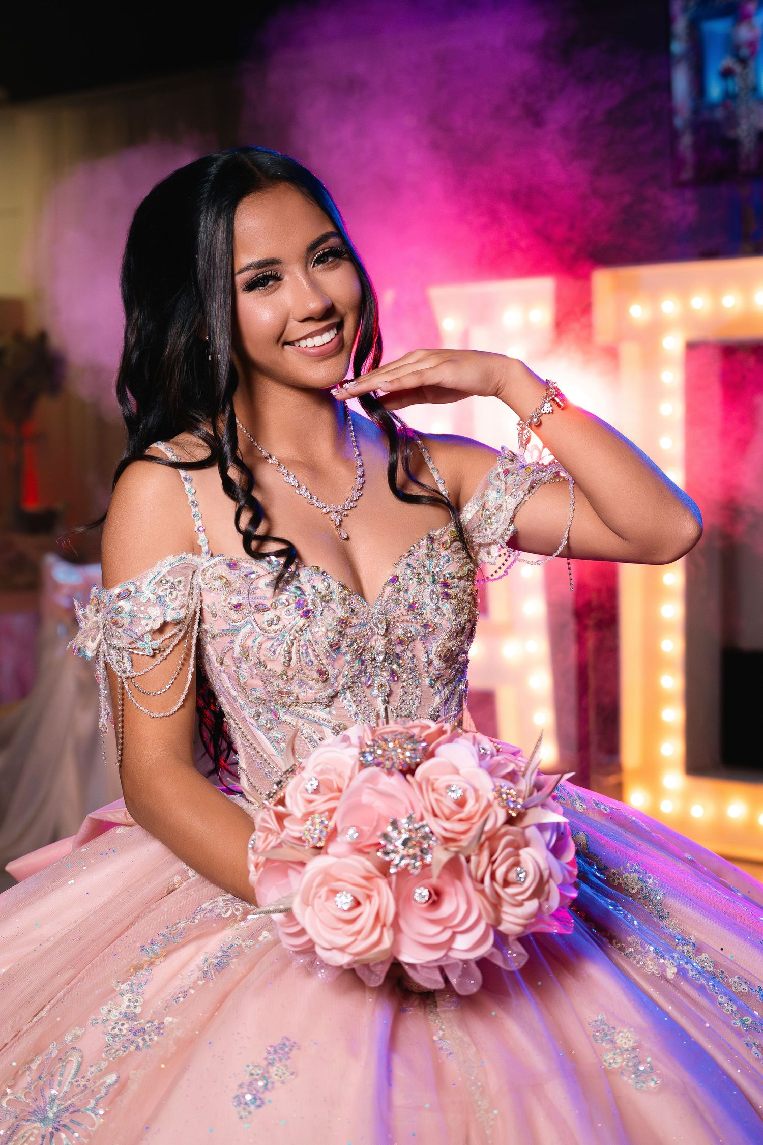 Young woman in pink quinceañera ball gown smiles holding floral bouquet at party.