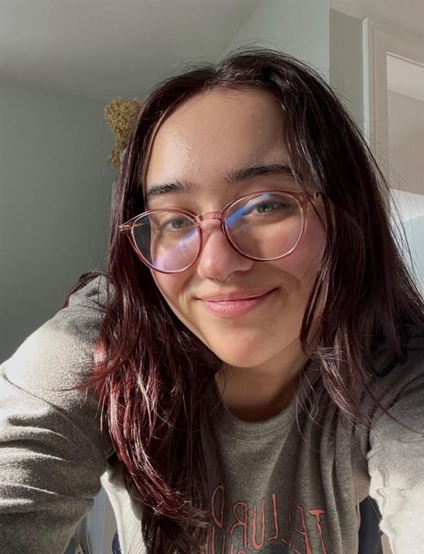 Young woman with dark red hair and round glasses smiling in a casual selfie photo.