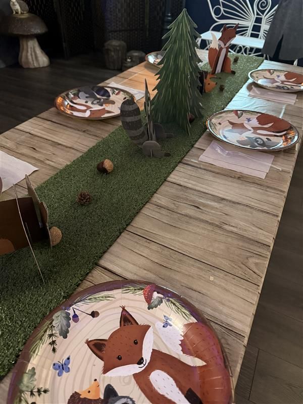 Woodland themed party table with fox plates, green grass runner, pine tree centerpiece, and acorn decorations on wood table.