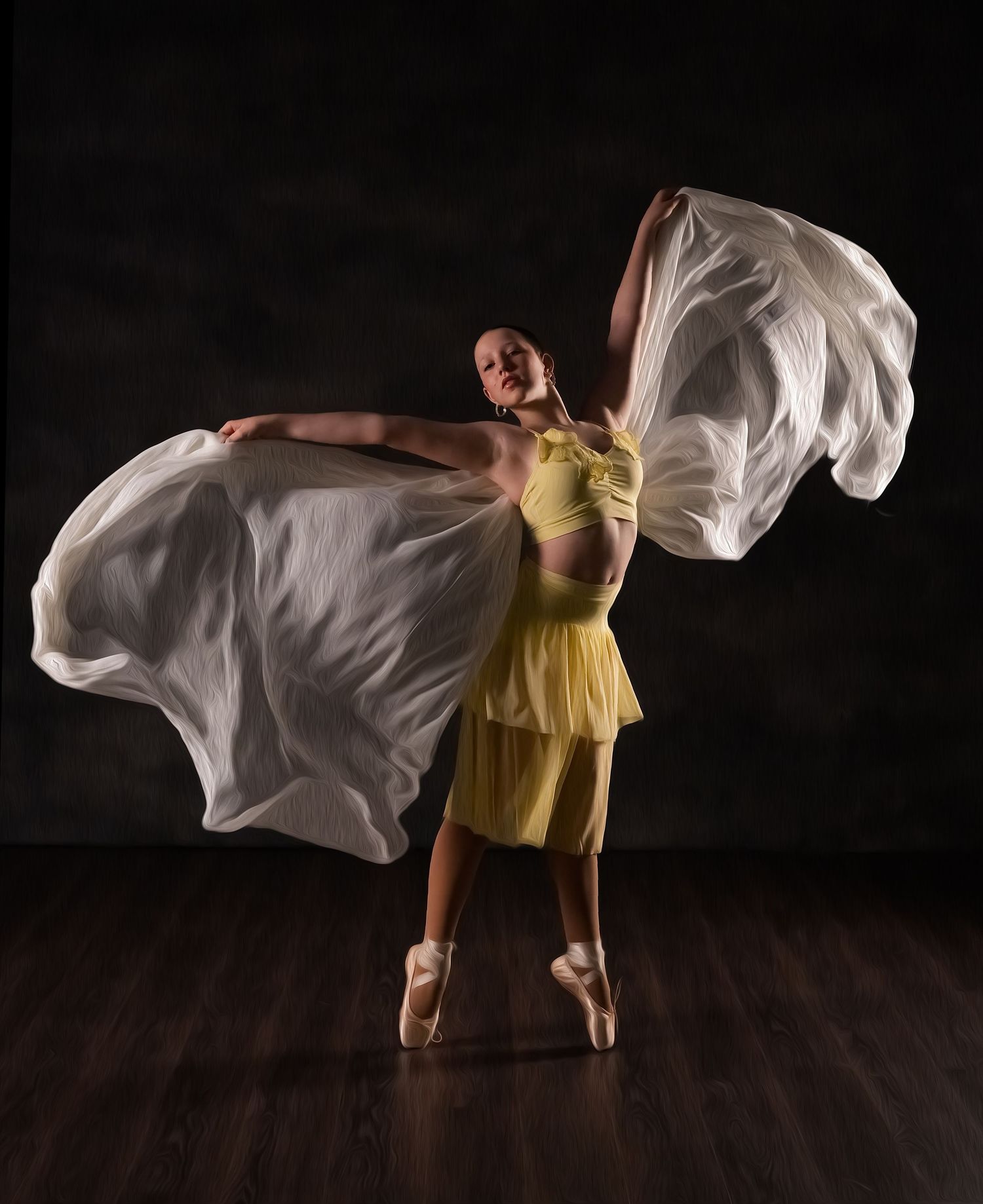 Ballerina in yellow dress on pointe shoes holds flowing white fabric wings against dark studio background.