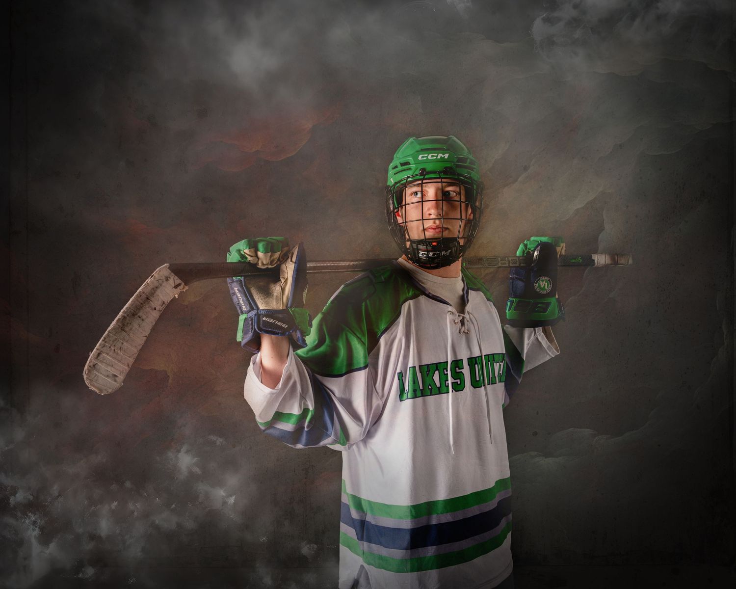Young hockey player in green helmet and white jersey poses with stick across shoulders against dramatic smoky background.