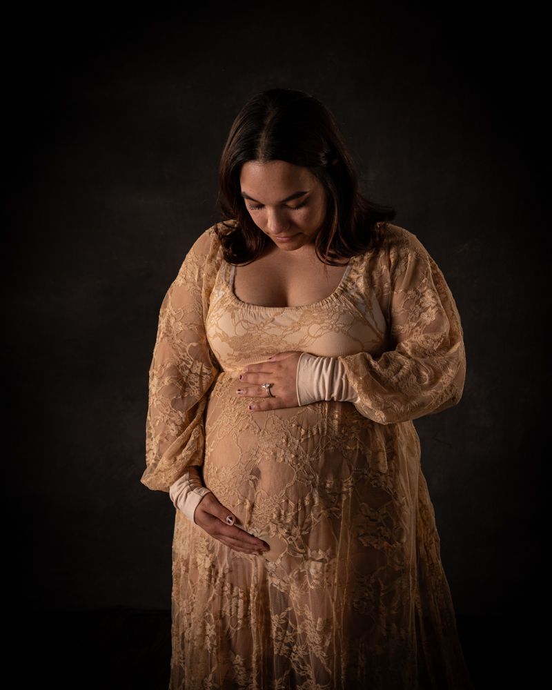 Pregnant woman in gold lace robe cradling baby bump against dark backdrop.