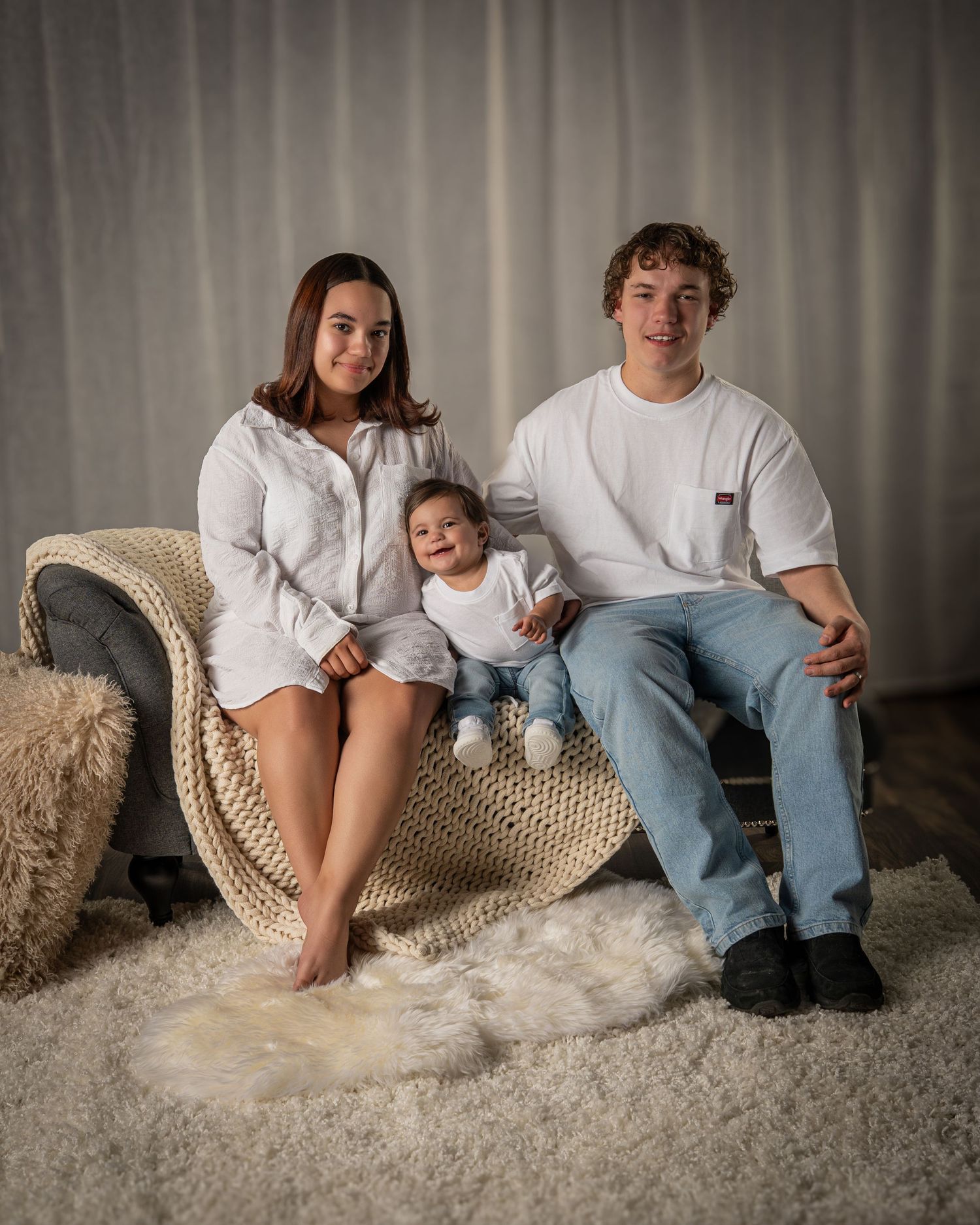 A smiling young family with an infant poses on a chaise lounge in a white studio.
