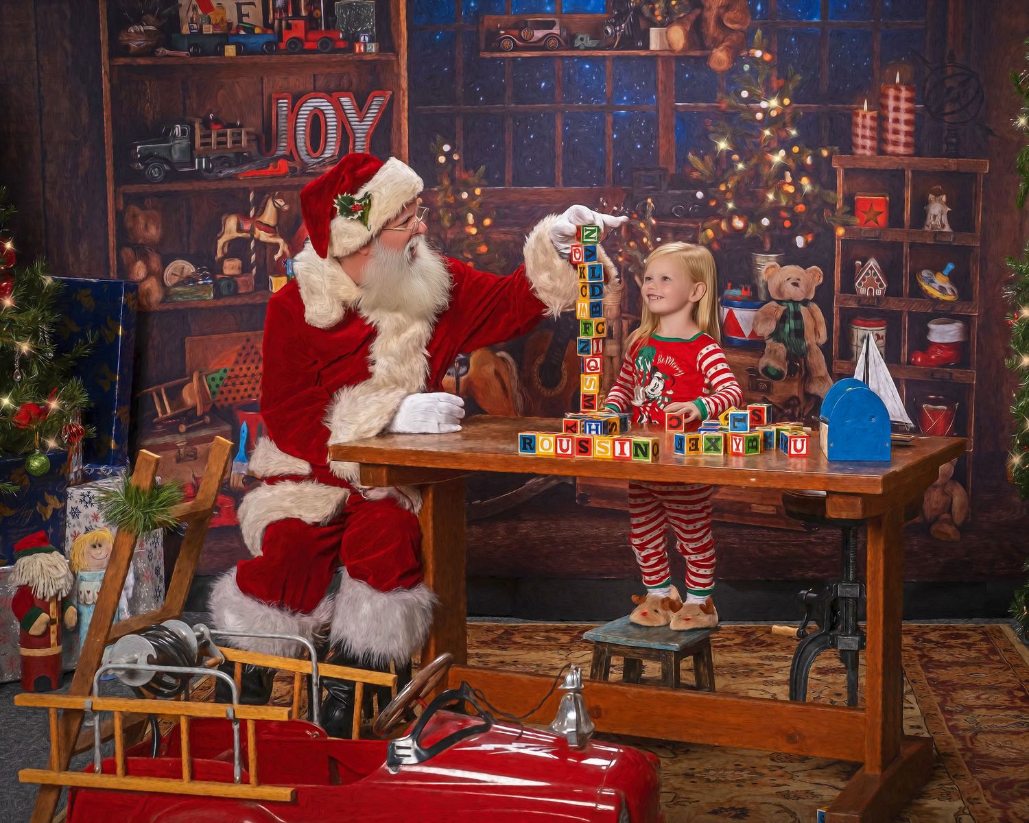 Santa Claus plays with colorful alphabet blocks with a young girl in red striped pajamas in a festive toy workshop.