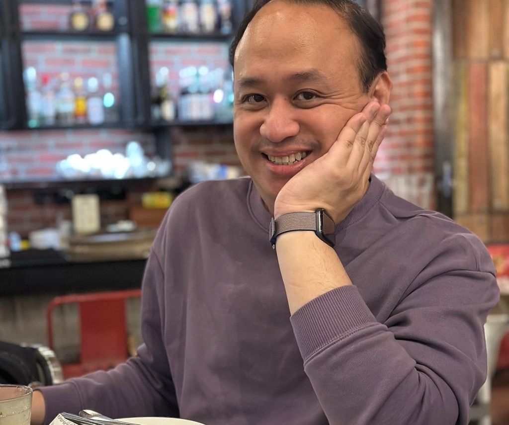 Smiling man resting chin on hand at a restaurant bar, wearing a purple shirt and smartwatch.