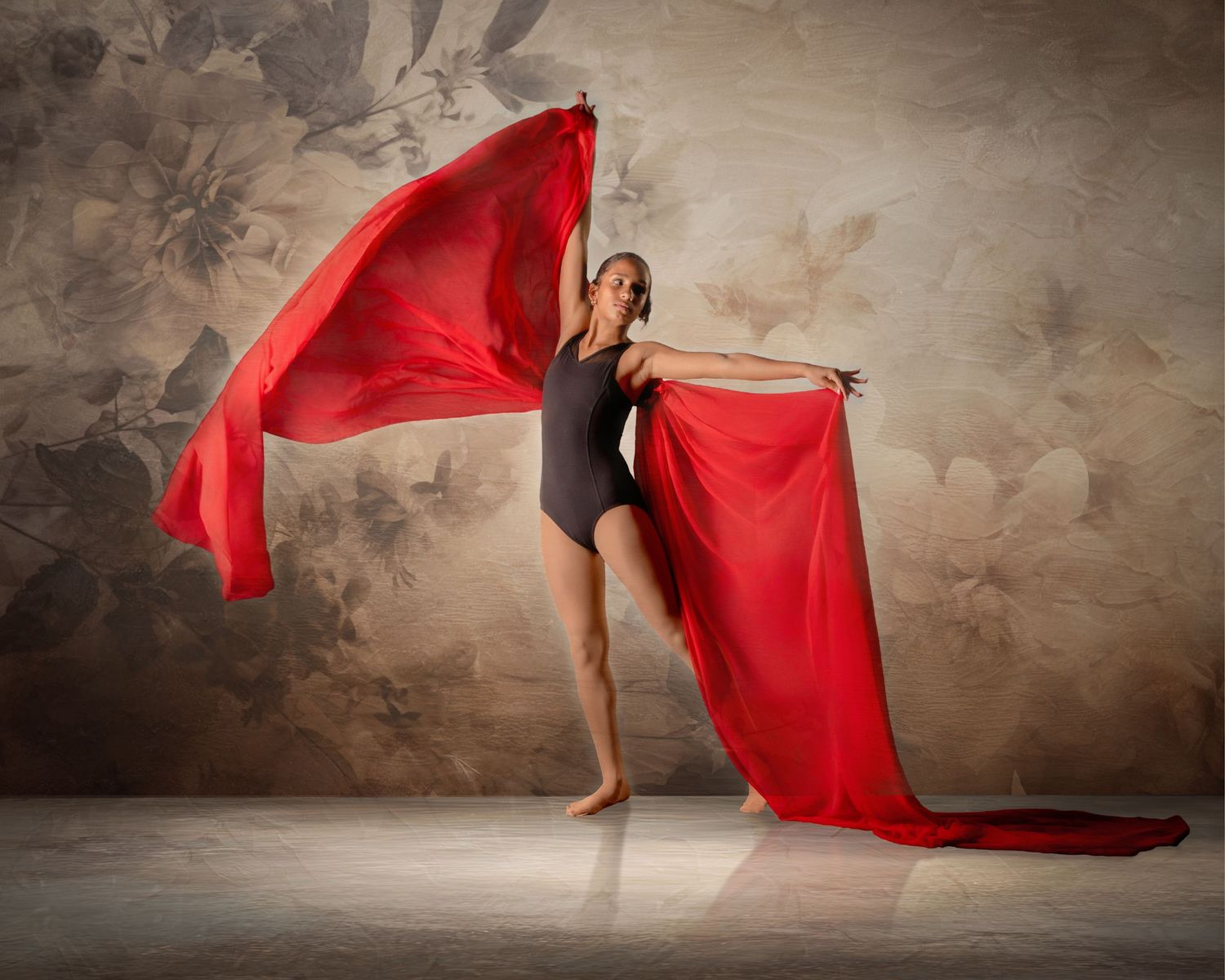 A graceful dancer in a black leotard elegantly poses while holding flowing red fabric against a floral backdrop.