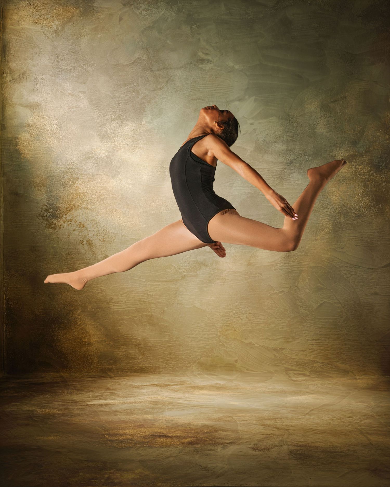 A dancer leaps gracefully in a black leotard against a golden textured backdrop, legs split mid-air in a dynamic pose.