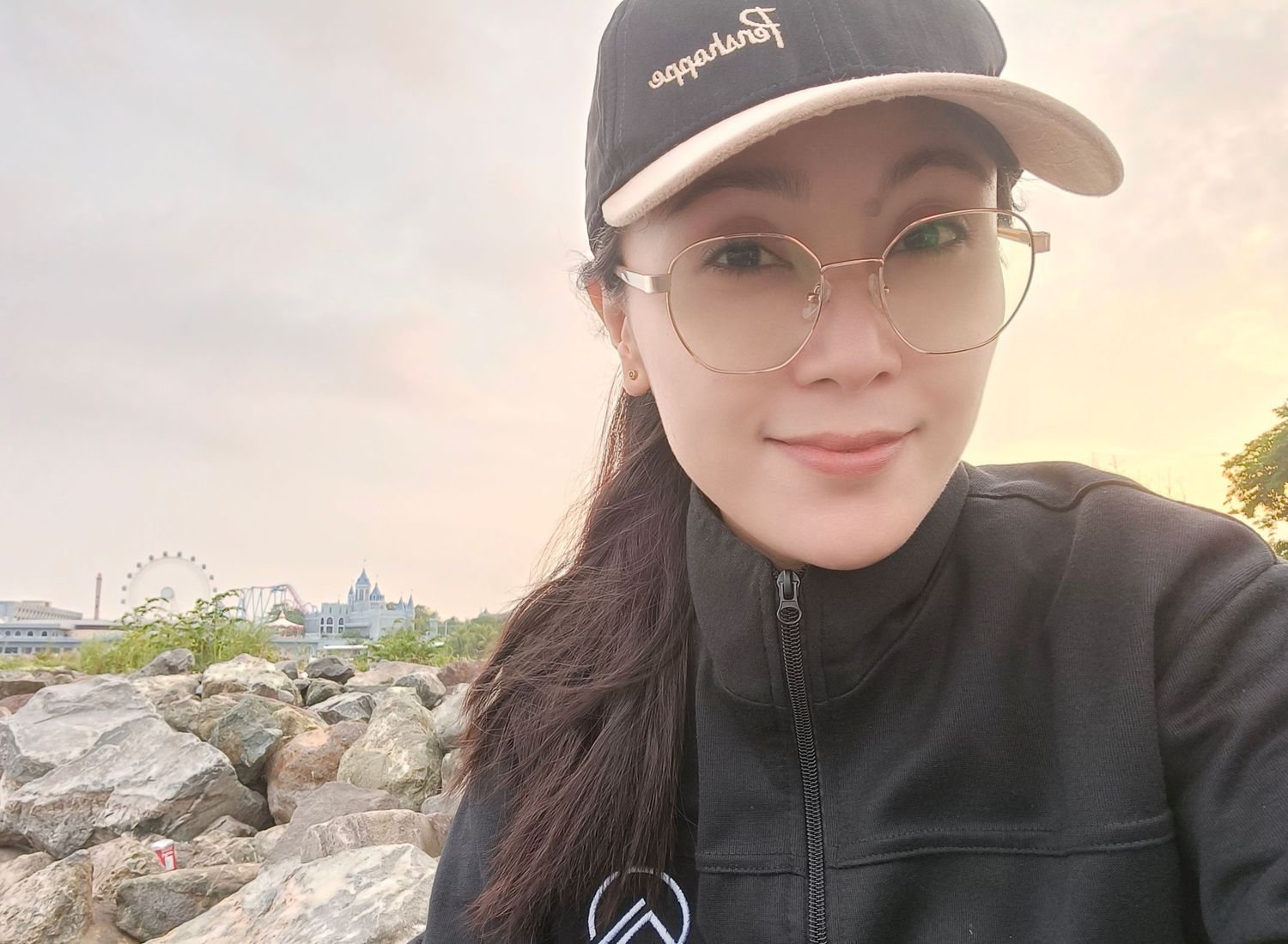 A young woman wearing glasses and a black cap poses for a selfie near a rocky waterfront with a Ferris wheel at sunset.