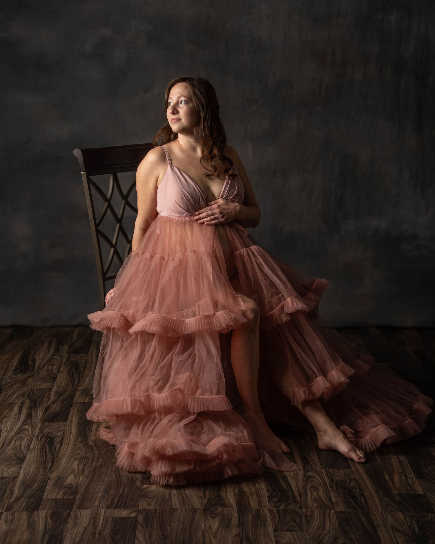 Pregnant woman in layered pink tulle gown seated on chair in studio.