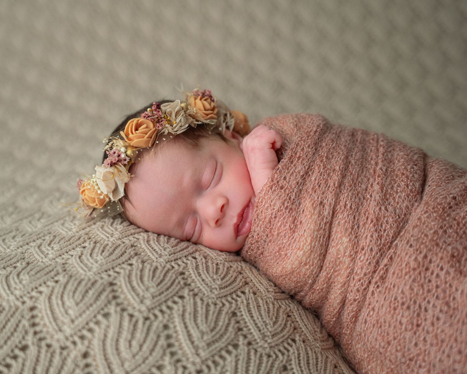 Sleeping newborn baby wrapped in pink swaddle with floral crown on textured knit blanket.