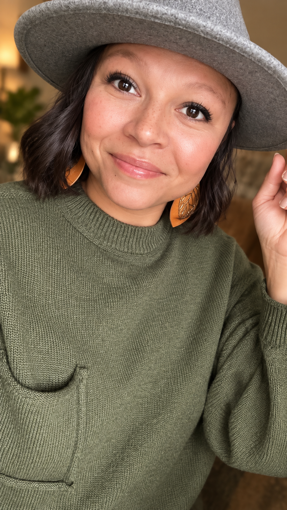 Woman in olive green sweater and gray wide-brim hat wearing brown leather earrings, smiling warmly indoors.