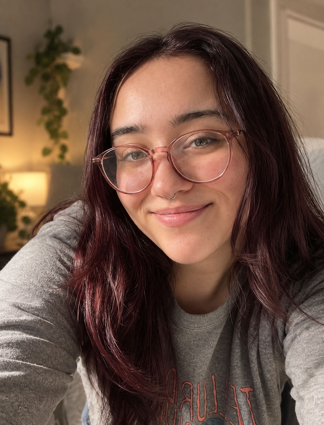 Young woman with long dark hair and rose-gold glasses smiling warmly in a cozy room selfie.
