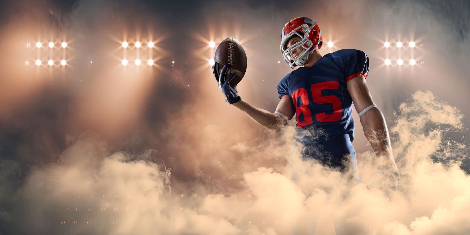 American football player in blue and red uniform holding ball aloft in dramatic smoky stadium lighting.