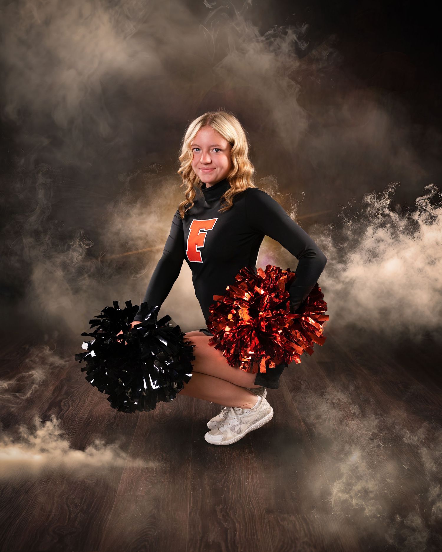 Young cheerleader in black and orange uniform poses with red and black pom-poms against a dramatic smoky background.