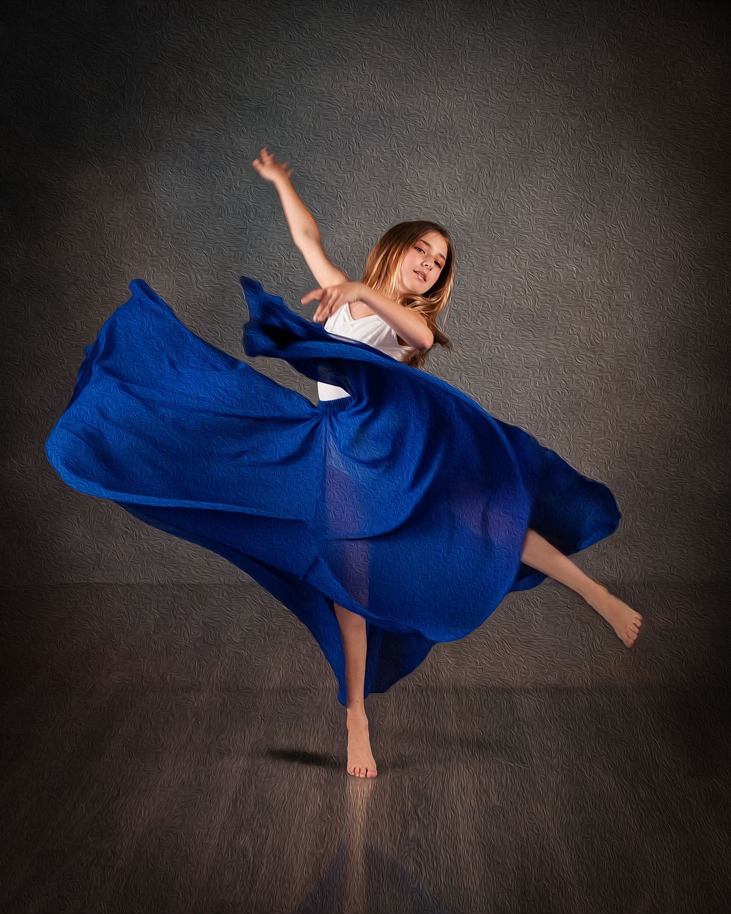 A young dancer leaps gracefully in a flowing royal blue skirt against a dark studio backdrop.