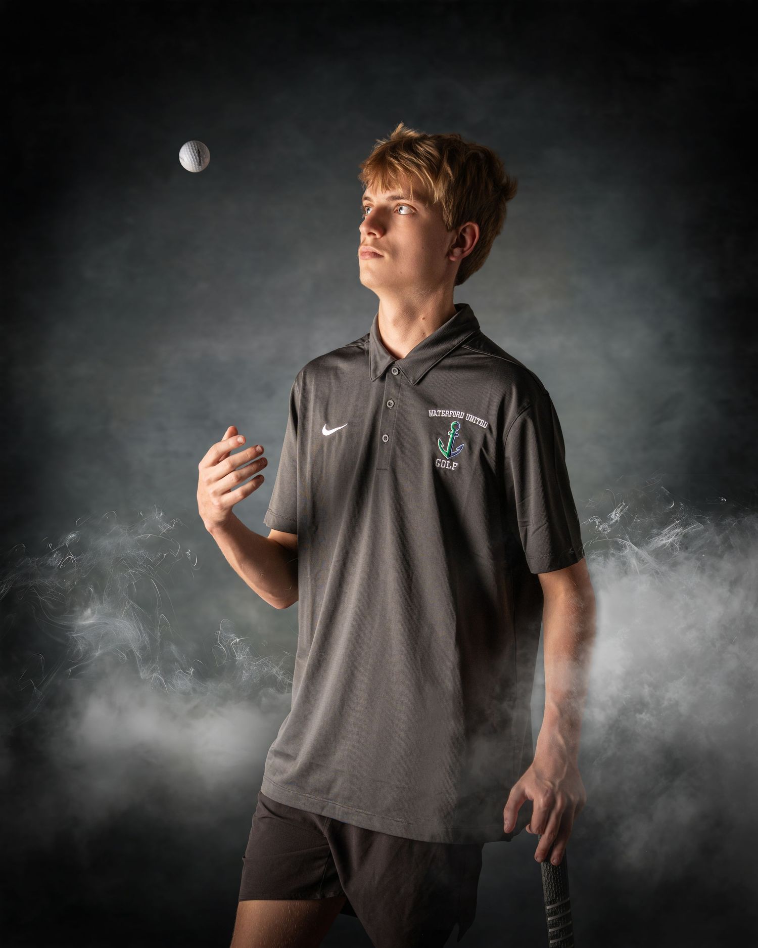 Young male golfer in gray Nike polo holds a club while tossing a golf ball against a dramatic smoky background.