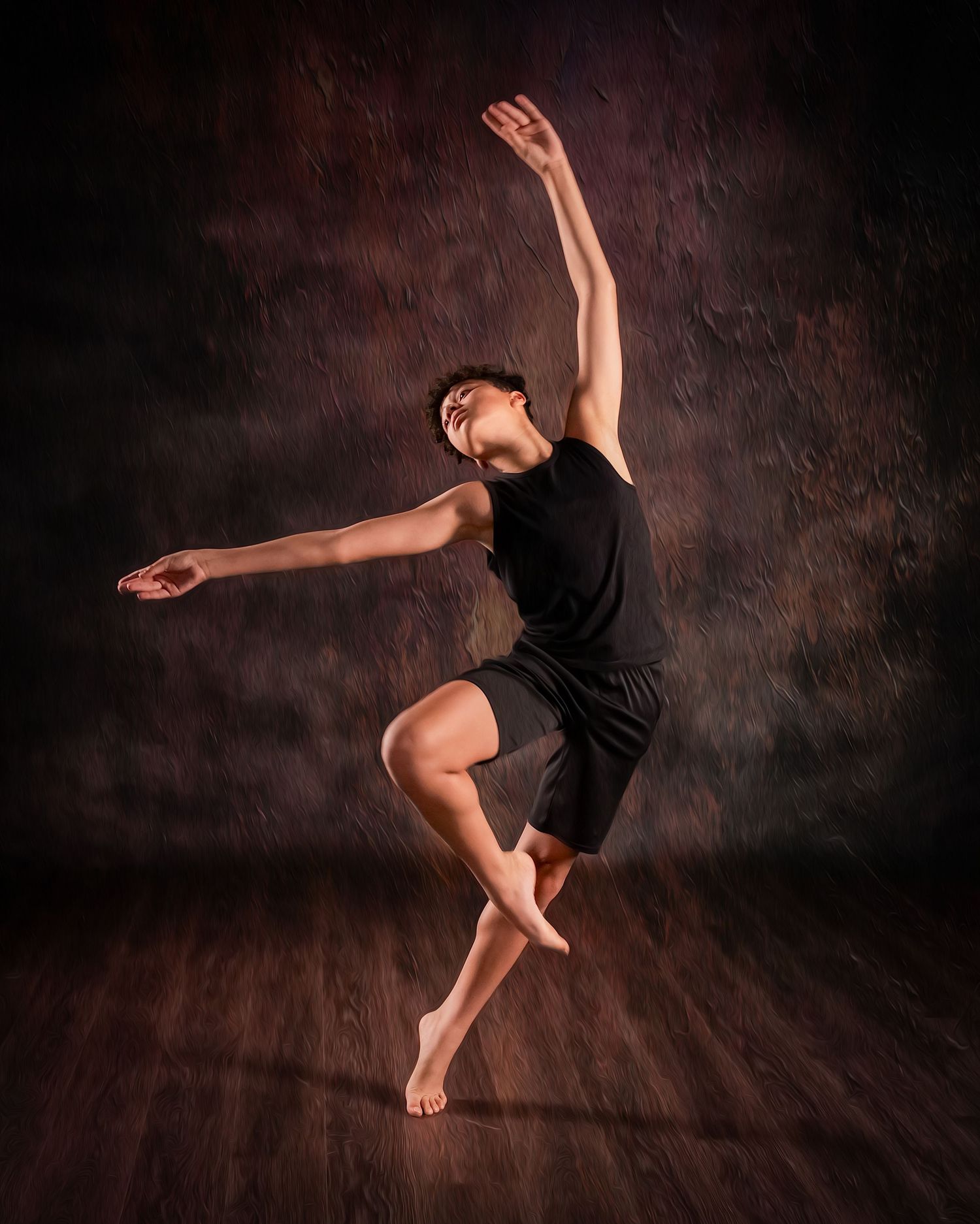 Male dancer leaping gracefully in black outfit against dark studio backdrop, one arm raised overhead on hardwood floor.