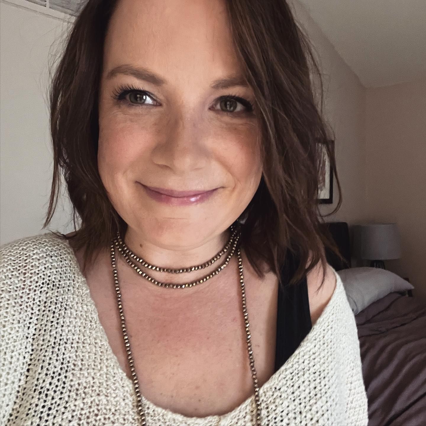 Smiling brunette woman taking a selfie wearing a cream cardigan and layered necklaces.