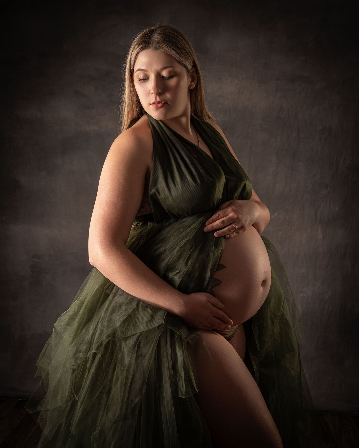 A pregnant woman in an olive green tulle gown poses against a dark studio backdrop.