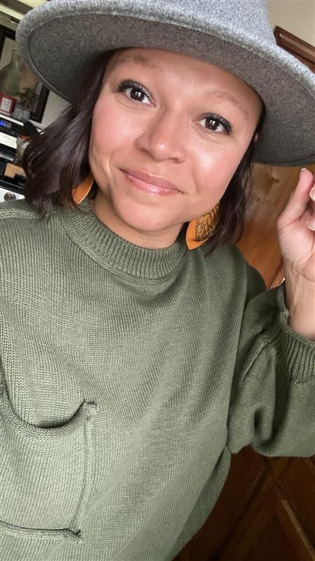 Smiling woman in gray hat and green sweater with orange earrings selfie.