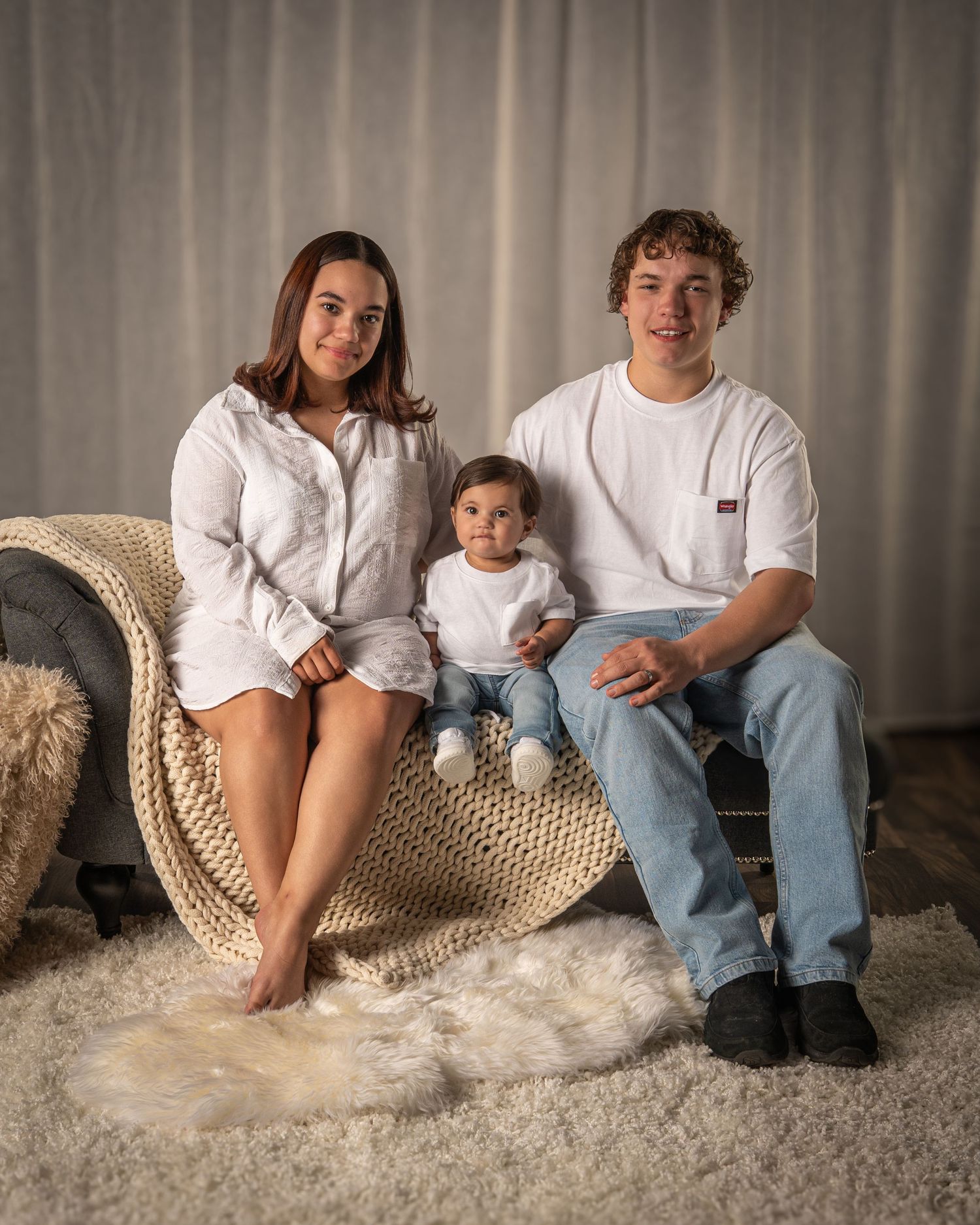 A young couple and their baby sit together on a chaise lounge in a bright studio setting.