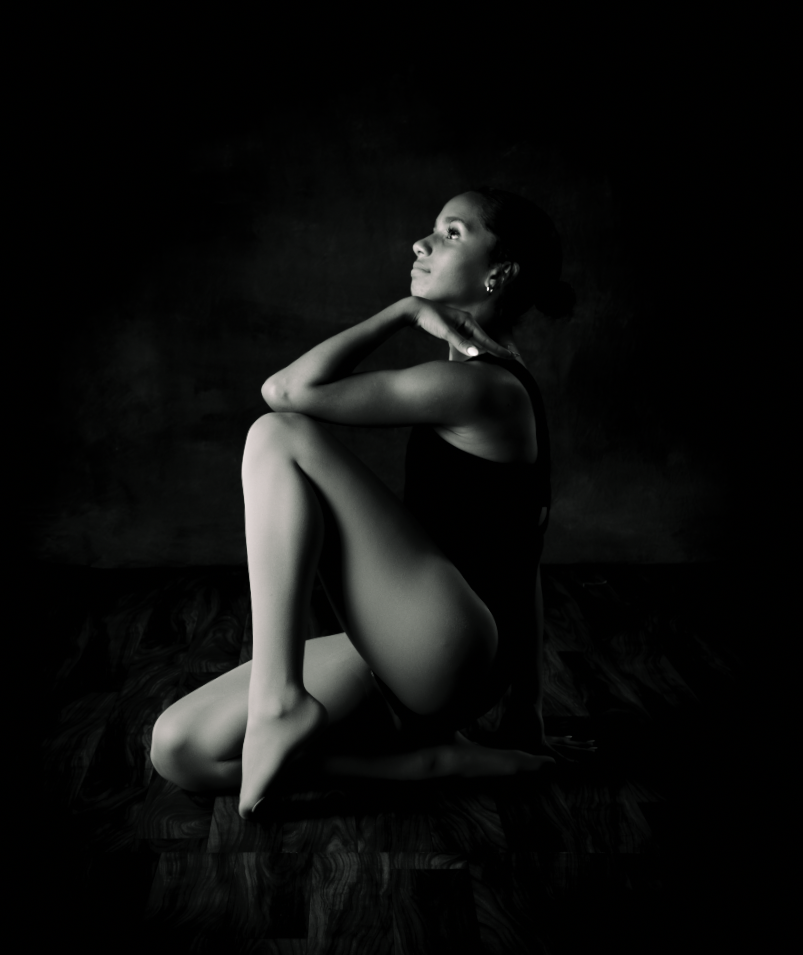 Black and white fine art photo of a woman sitting gracefully with knees drawn up, gazing upward in dramatic studio lighting.