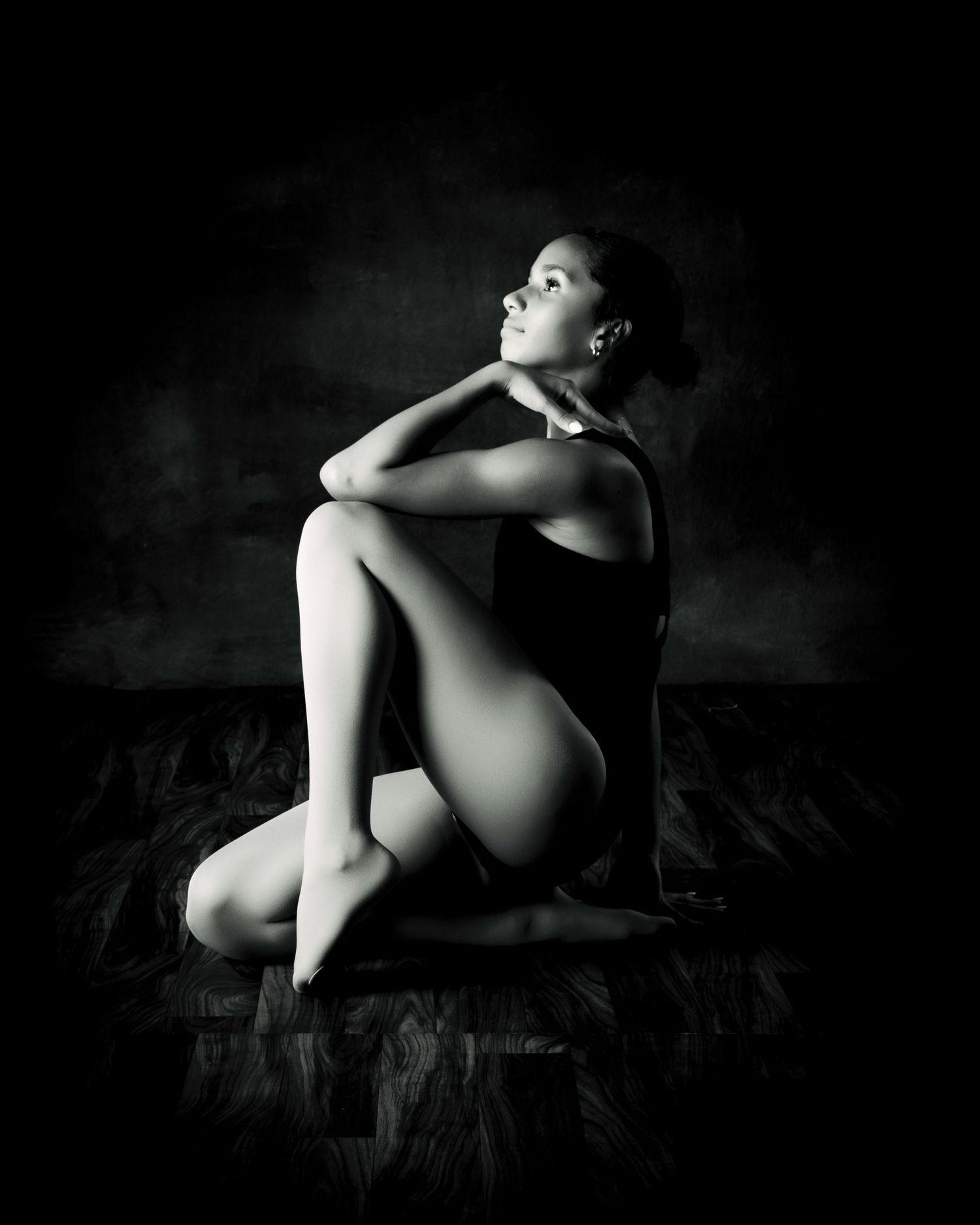 Black and white fine art photo of a young woman in a leotard seated gracefully on a dark floor, gazing upward.