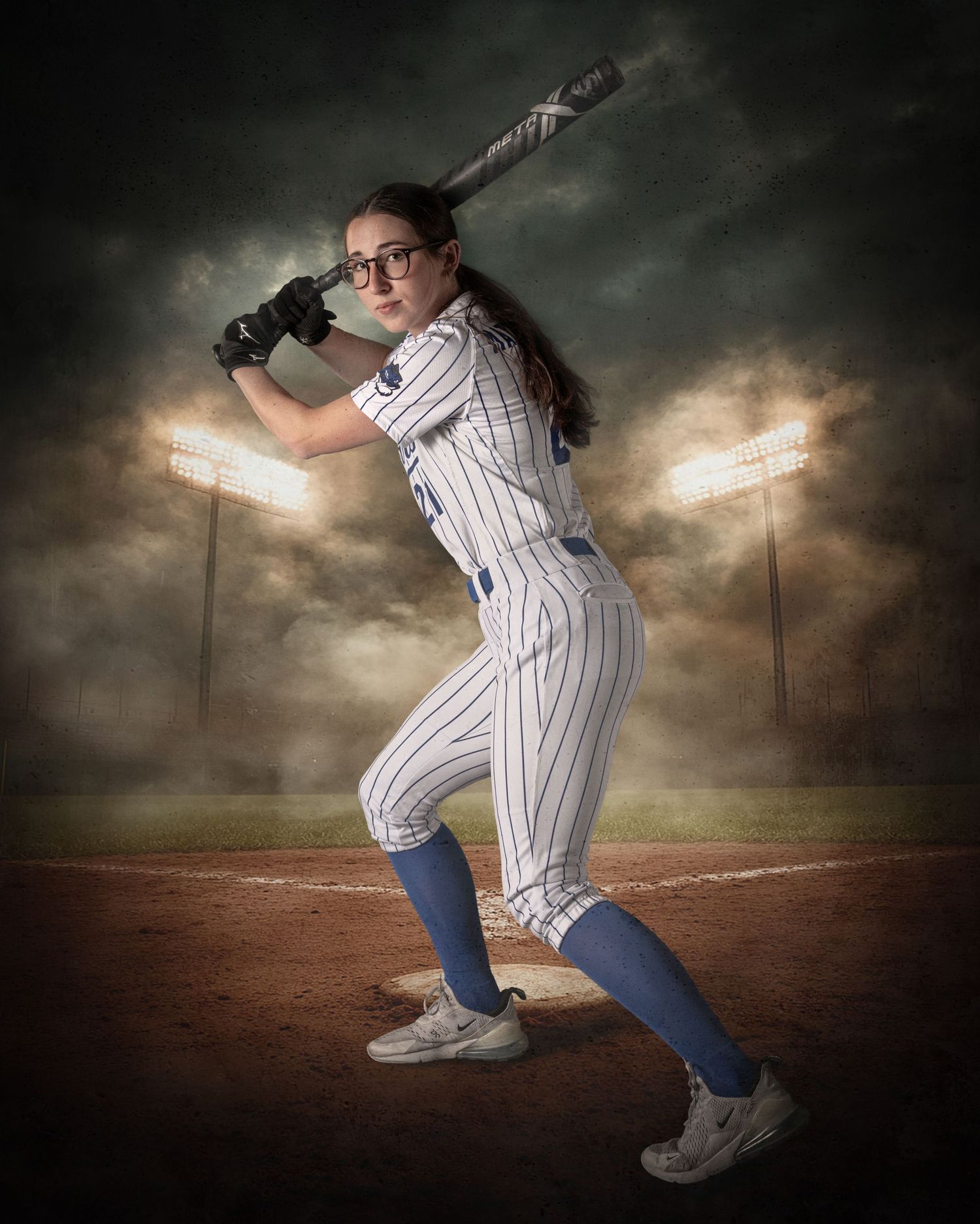 Female softball player in white pinstripe uniform poses in batting stance under stadium lights on a dramatic cloudy field.