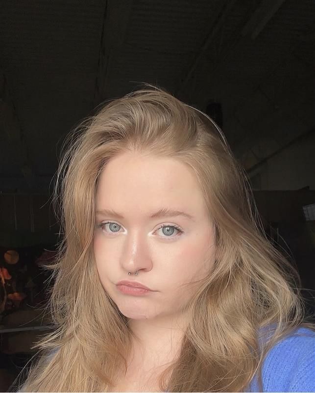 Young blonde woman with blue eyes taking a selfie wearing a blue top with hair down.