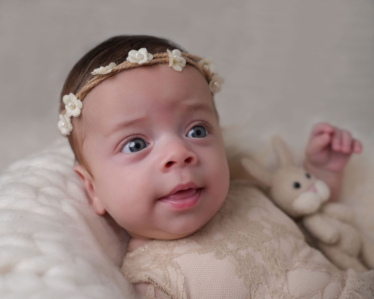 Adorable baby with blue eyes wearing a delicate floral headband lying on a soft blanket with a bunny toy.