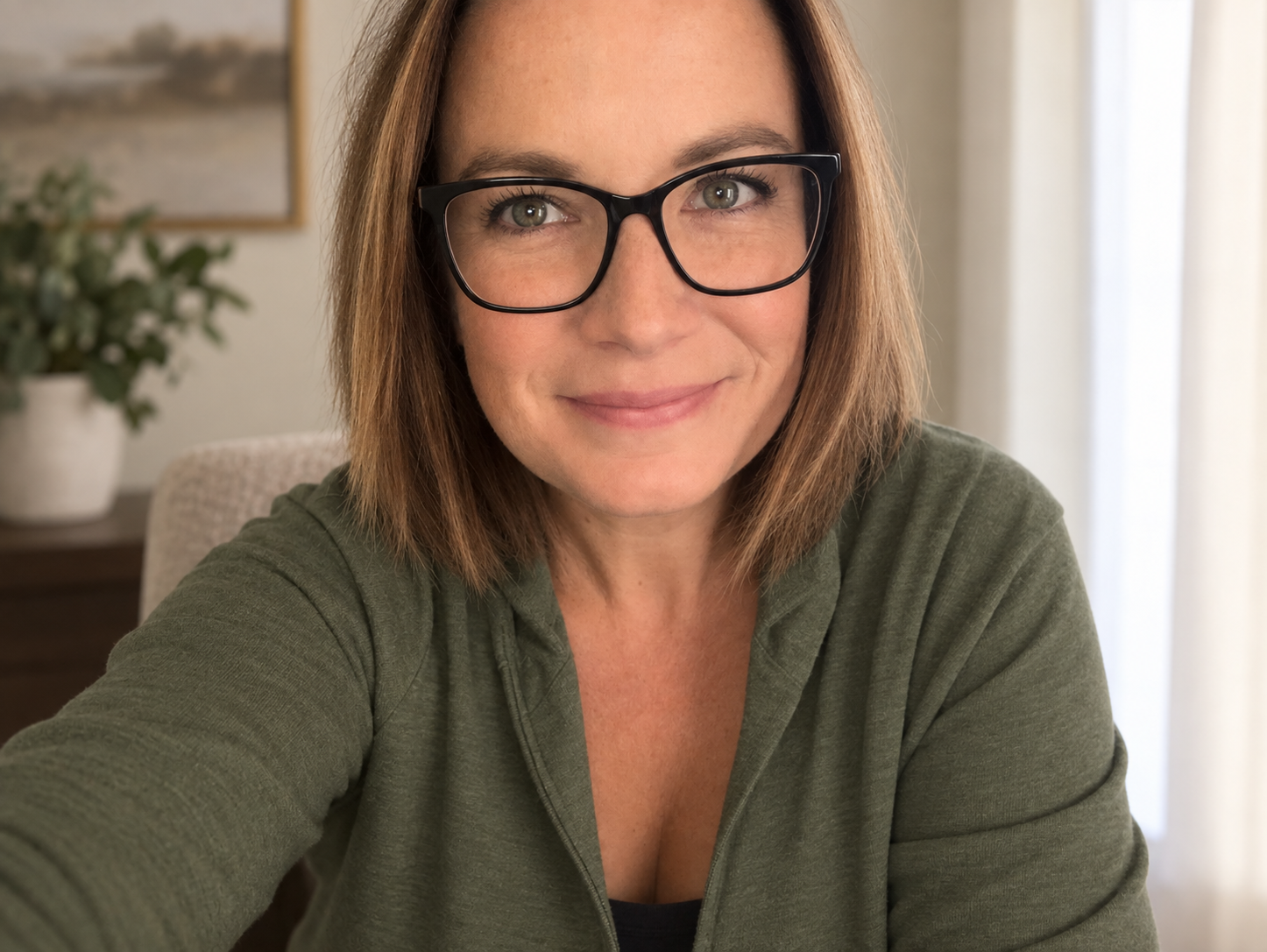 Woman with glasses and shoulder-length brown hair smiling in a selfie, wearing a green cardigan indoors.