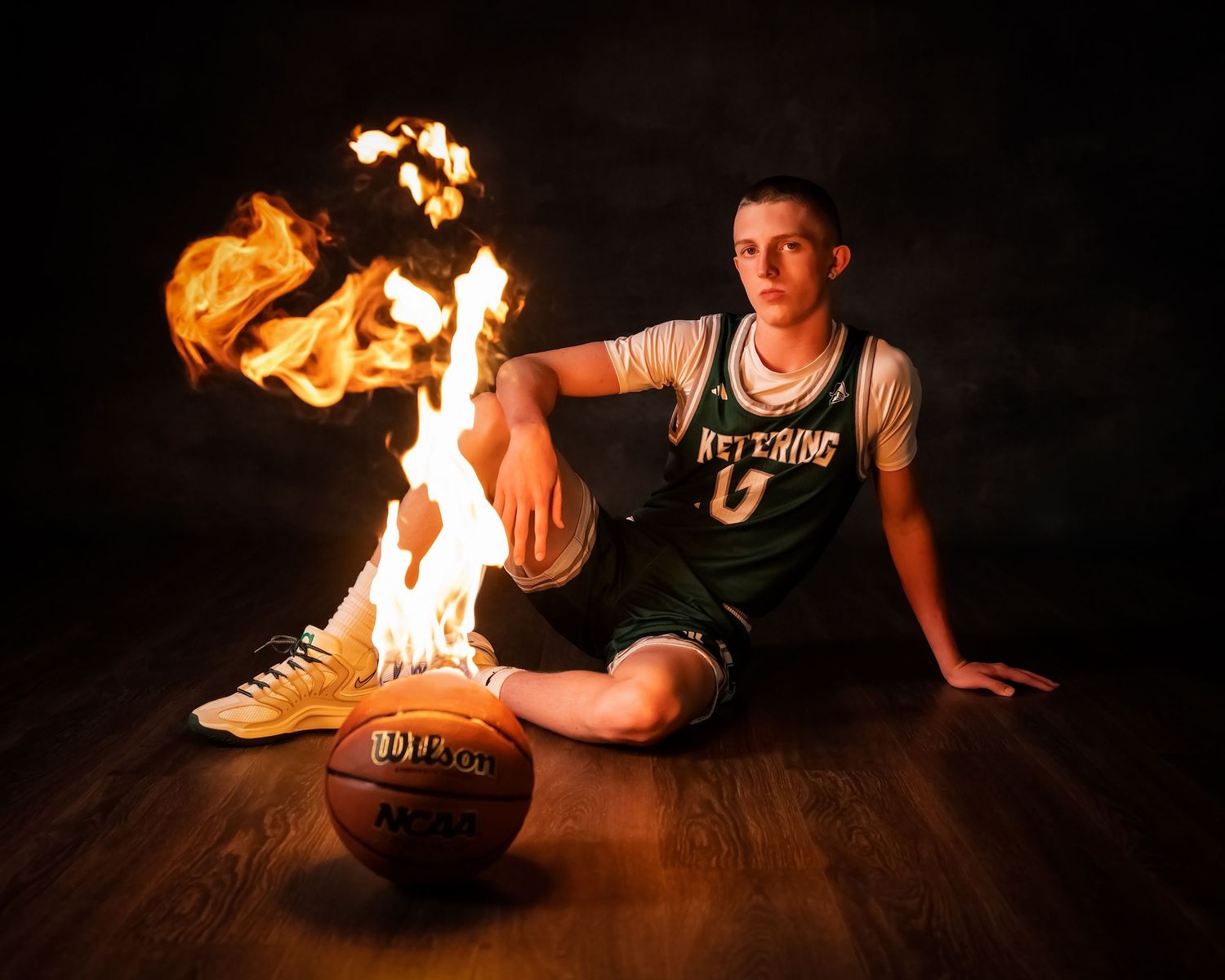 Basketball player in green Kettering jersey sits on court floor beside flaming sneakers with a basketball nearby.