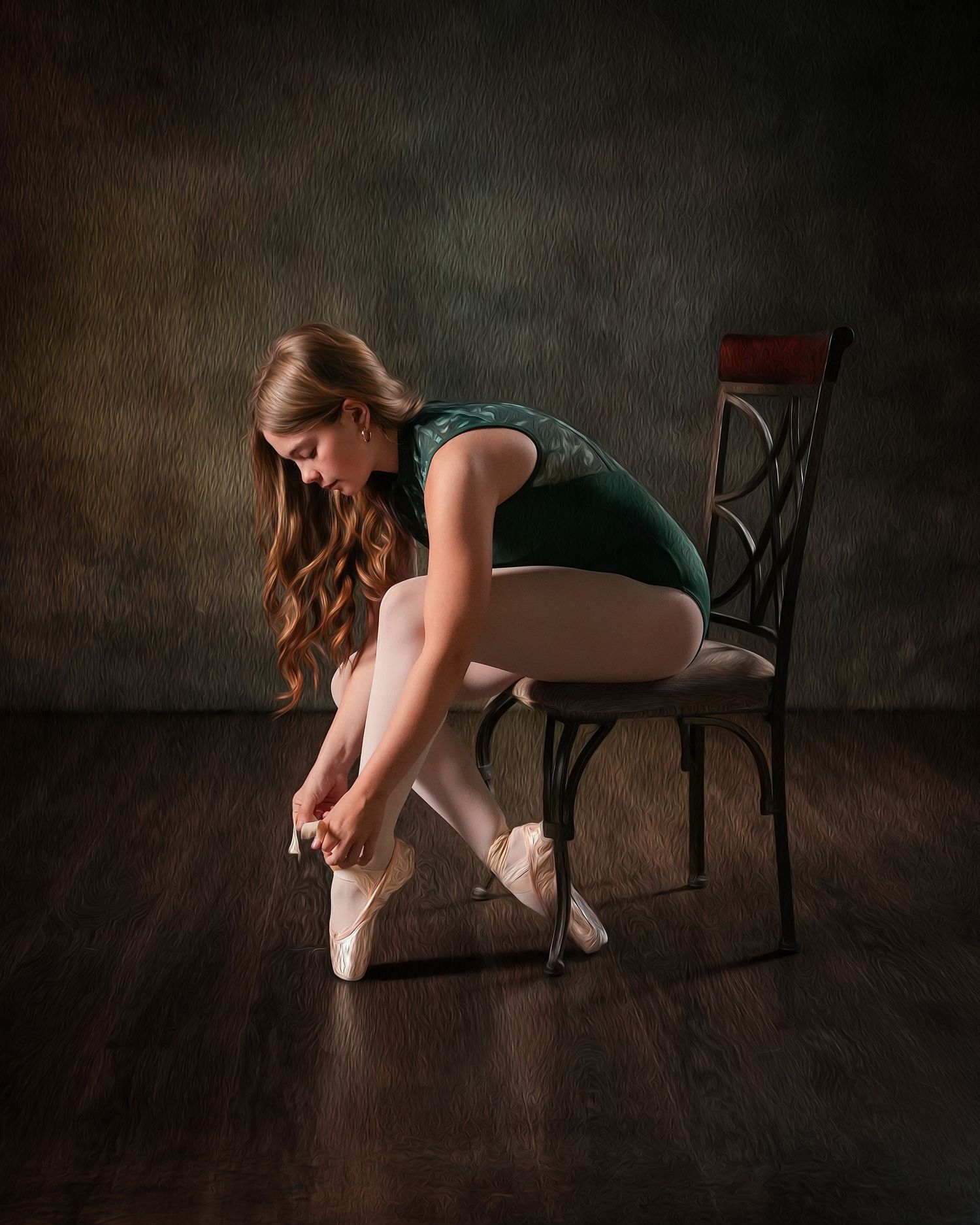 A ballerina in a green leotard sits on a chair tying her pointe shoes on a dark wood floor.