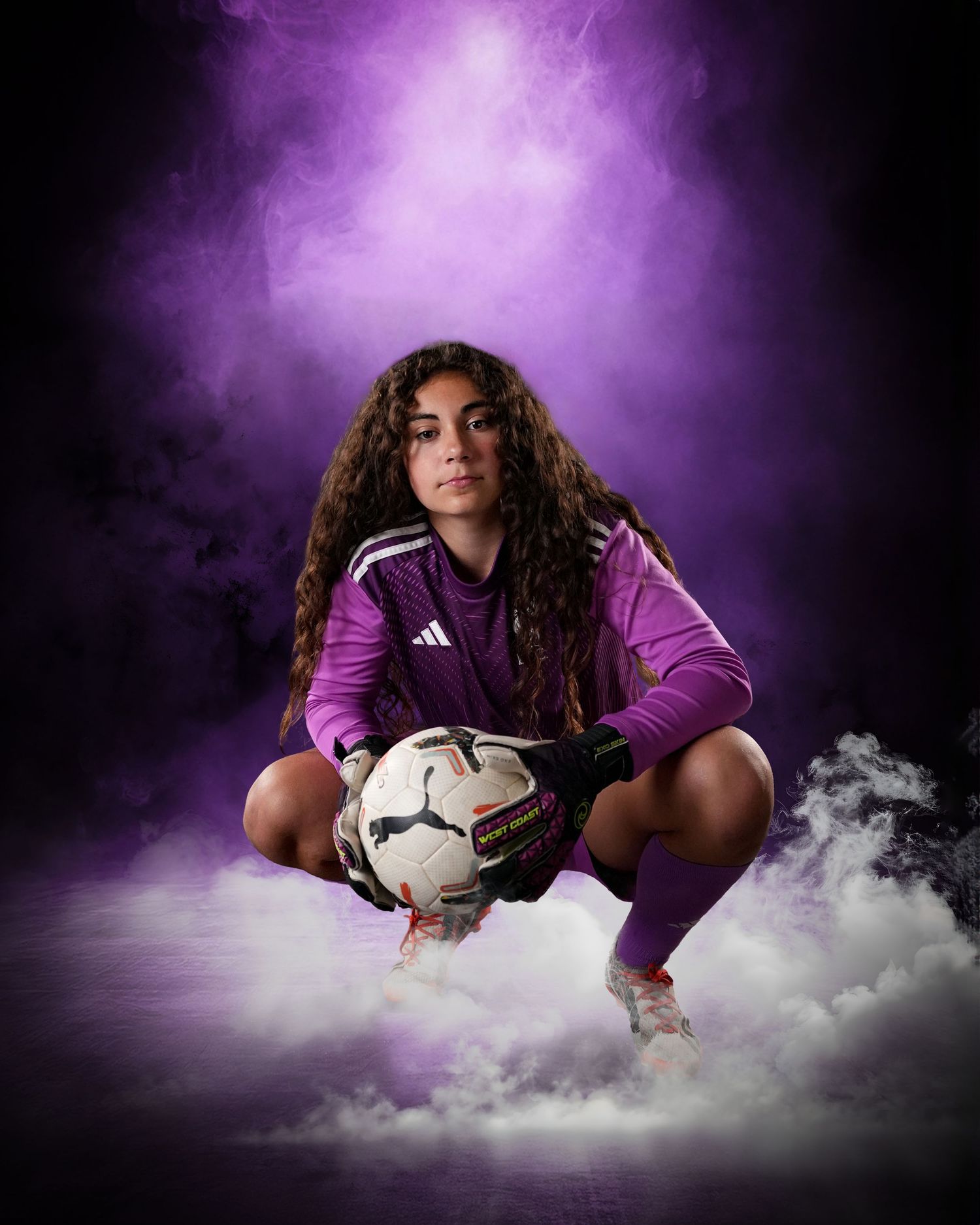 Female soccer goalkeeper in purple Adidas kit crouching with Puma ball amid dramatic purple smoke.