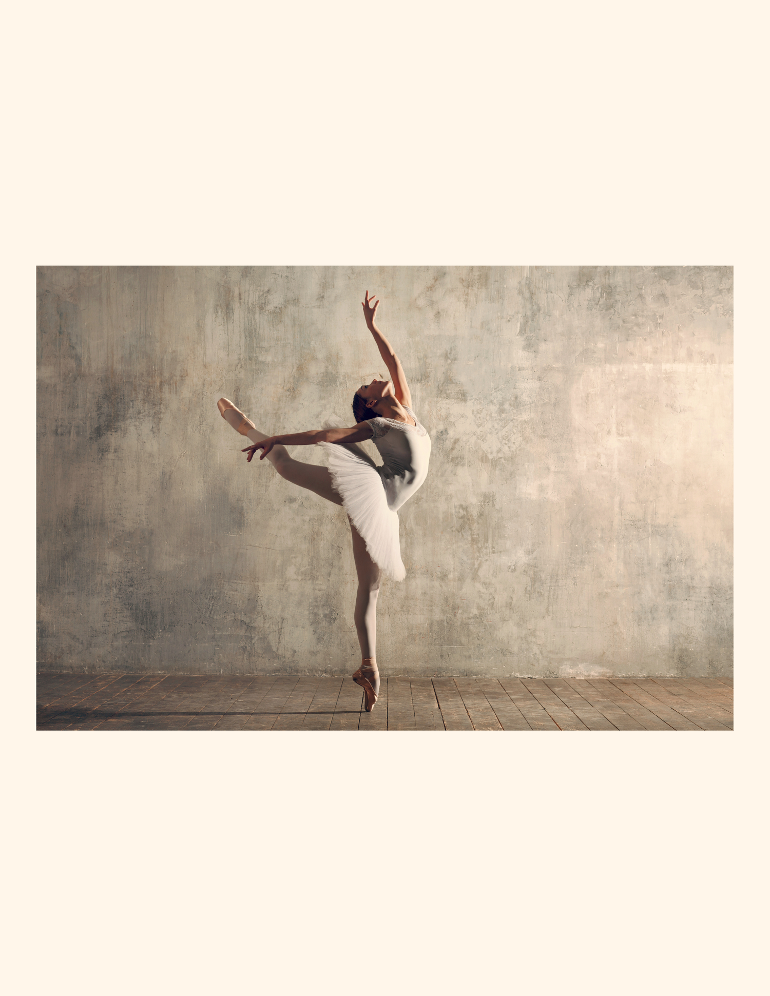 A ballet dancer performs an elegant arabesque pose on pointe in a gray studio.