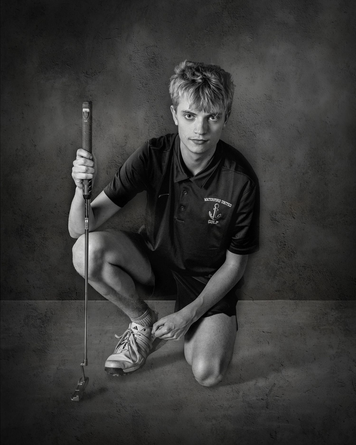 Black and white portrait of a young male golfer kneeling while holding a golf club in a studio setting.