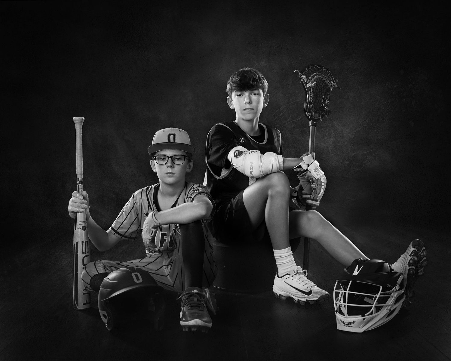 Black and white photo of two boys posing with sports equipment, one holding a baseball bat and the other a lacrosse stick.