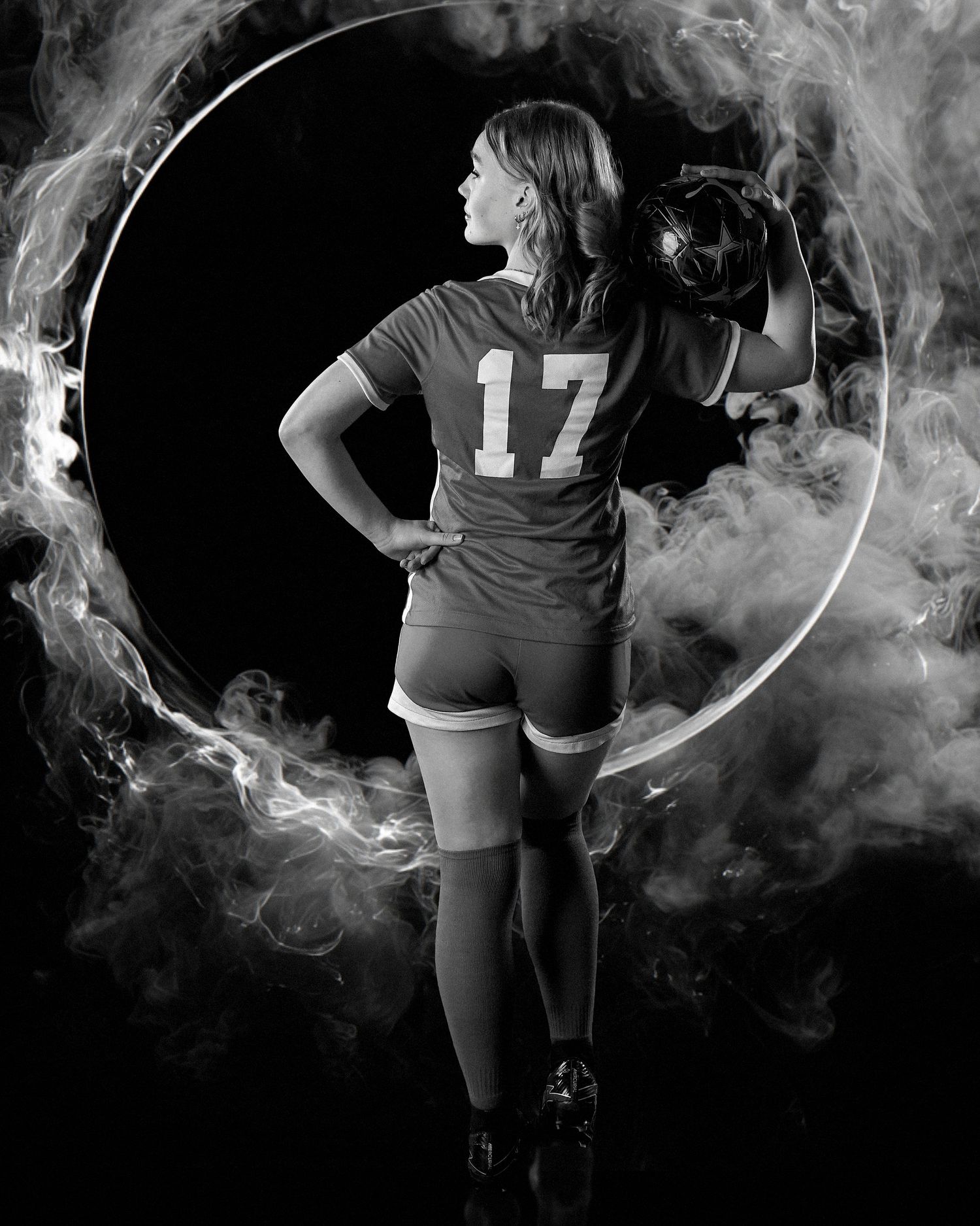 Black and white photo of female athlete in jersey number 17 holding helmet, surrounded by dramatic smoke ring effect.