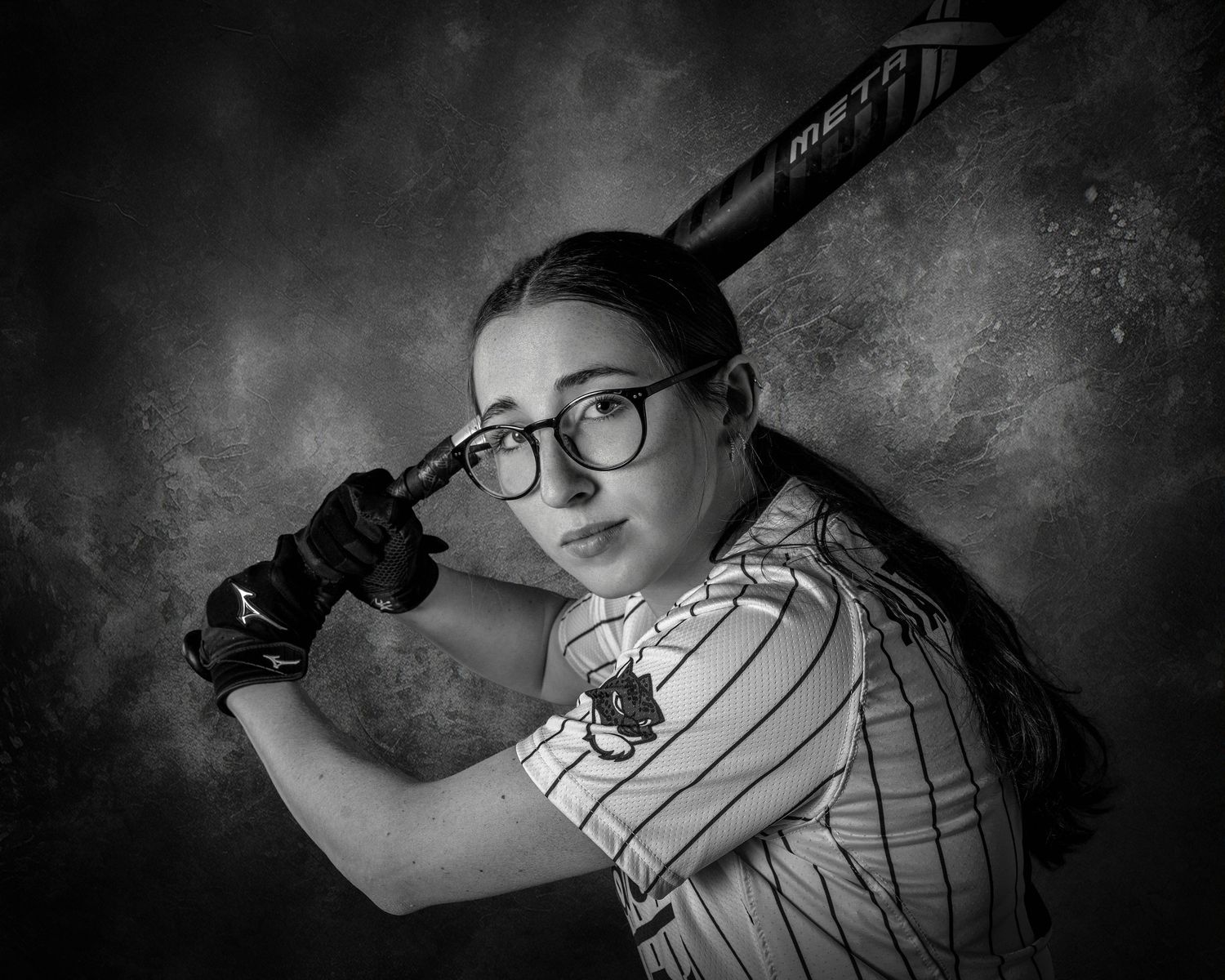 Black and white portrait of a young softball player in batting stance, wearing glasses and pinstripe jersey, holding a Meta bat.